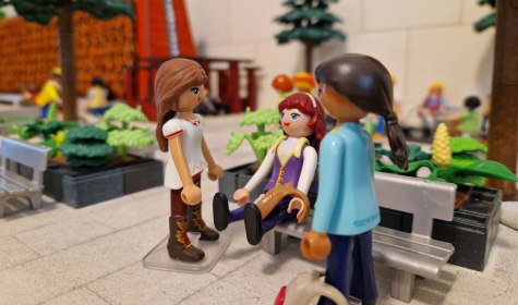 Bad Salzufler Gebäude und Bauwerke wie das Gradierwerk am Schliepsteiner Tor (Hintergrund) erwecken die Playmobil-Sammler Nicole und Michael Post im Kleinformat mit der Sonderausstellung „Nachgebaut! Bad Salzuflen aus Playmobil“ ab dem 06. November 2026 in der Wandelhalle zum Leben, © Stadt Bad Salzuflen Drei Playmobil-Figuren in einem Park mit Bänken und Bäumen, eine sitzt auf der Bank, zwei stehen davor