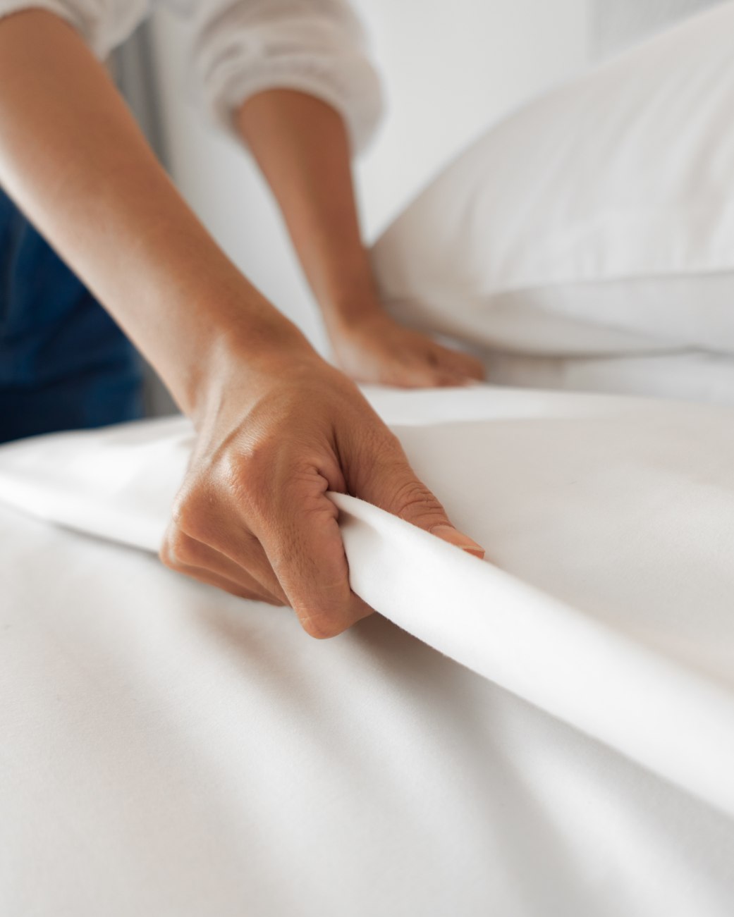 Eine Hand bereitet sorgfältig ein mit weißer Bettwäsche bezogenes Doppelbett im Hotelzimmer vor, © iStock-1194628093