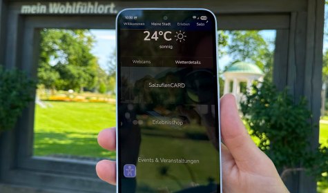 Person hält Smartphone mit dem Startbildschirm der MeinS App in der Hand. Im Hintergrund sind der Leopoldsprudel und der Kurpark der Stadt Bad Salzuflen zu sehen, © Stadt Bad Salzuflen Person hält Smartphone mit dem Startbildschirm der MeinS App in der Hand. Im Hintergrund sind der Leopoldsprudel und der Kurpark der Stadt Bad Salzuflen zu sehen, © Stadt Bad Salzuflen