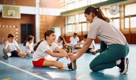 Auf dem Foto ist eine Turnhalle zu sehen. Im Vordergrund hockt eine junge Trainerin, die ein Kind dabei unterstützt sich zu dehnen., © stock.adobe.com - Drazen 