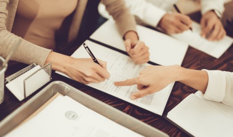 Auf dem Bild sind drei Personen zu sehen. Sie sitzen um einen Schreibtisch und haben Unterlagen auf dem Tisch ausgebreitet. Eine Person ist dabei ihre Unterschrift zu setzen. Es wird ein Vertrag über eine außergerichtliche Beilegung einer Streitigkeit aufgesetzt., © VadimGuzhva - stock.adobe.com