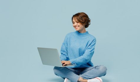 Frau sitzt im Schneidersitz auf dem Boden und schaut auf das aufgeklappte Notebook, das auf ihrem Scho&szlig; liegt, &copy; Adobe Stock
