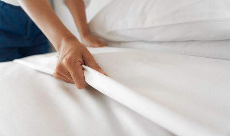 Eine Hand bereitet sorgfältig ein mit weißer Bettwäsche bezogenes Doppelbett im Hotelzimmer vor, © iStock-1194628093