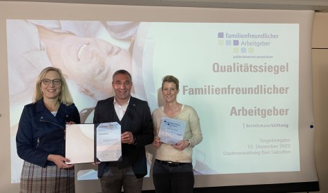 3 Personen stehen vor einer Leinwand, auf der eine Präsentation zur Zertifizierung als &quot;familienfreundlicher Arbeitgeber&quot; der Bertelsmann-Stiftung angezeigt sind, die Personen halten eine Urkunde und eine Plakette in den Händen. Die Personen sind von links nach rechts Prüferin Dr. Alexandra Heinzelmann, Bürgermeister Dirk Tolkemitt und Erste Beigeordnete Melanie Koring, © Stadt Bad Salzuflen
