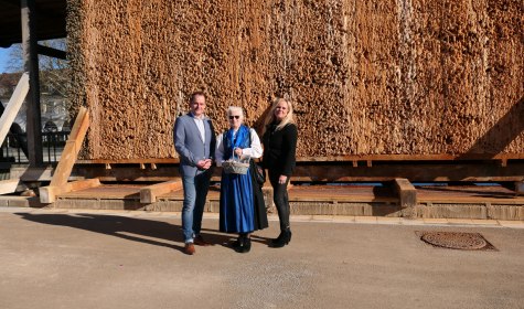Zu sehen sind drei Personen, sie stehen vor einem Gradierwerk in Bad Salzuflen. Die Person in der Mitte hält einen Präsentkorb in der Hand.