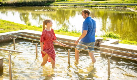 Eine Frau im roten Kleid und ein Mann in Shorts &amp; T-Shirt treten Wasser im Wassertretbecken auf der Kneipp-Insel im Kurpark Salzuflen