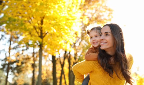 Das Bild zeigt eine Mutter mit ihrer Tochter auf dem Rücken, die draußen durch eine sonnige Herbstlandschaft laufen., © stock.adobe.com - New Africa