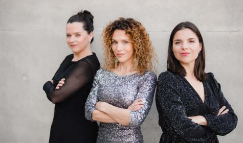 Erfrischend und originell ist der Exkurs durchs Leben der berühmten Sängerin Édith Piaf am 21. November 2026 mit der „Enttarnung einer Diva“ im Galasaal des Kurhauses in Bad Salzuflen, © Simon Detel Drei Frauen mit verschränkten Armen stehen vor einer hellen Wand, die mittlere trägt ein glitzerndes Kleid, die anderen zwei schwarze Kleider